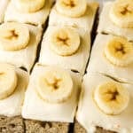 Square slices of banana cake with cream cheese frosting are all topped with a banana slice.
