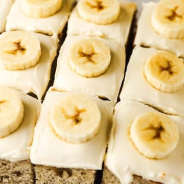 Square slices of banana cake with cream cheese frosting are all topped with a banana slice.