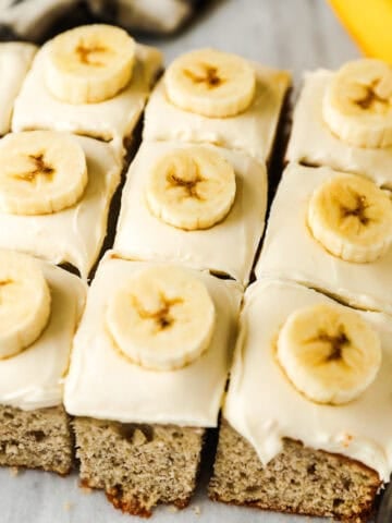Square slices of banana cake with cream cheese frosting are all topped with a banana slice.