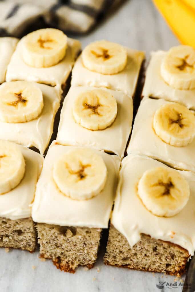 Square slices of banana cake with cream cheese frosting are all topped with a banana slice.