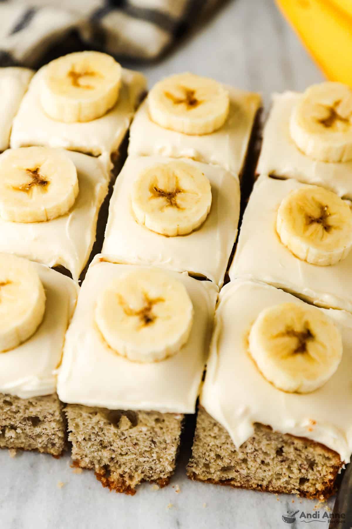 Square slices of banana cake with cream cheese frosting are all topped with a banana slice.