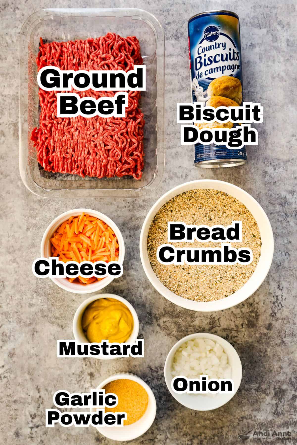 Ground beef and refrigerated biscuit dough in their packages, and cheese, bread crumbs, mustard, diced onion, and garlic powder in small white bowls.