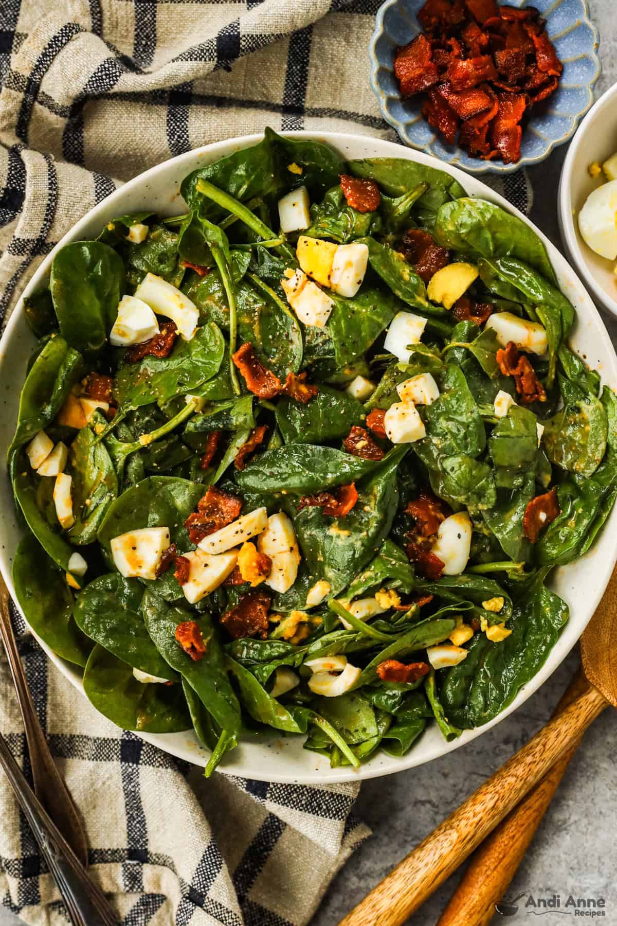 Classic spinach salad in a large white bowl and small side dishes with additional bacon pieces and chopped hard-boiled egg.