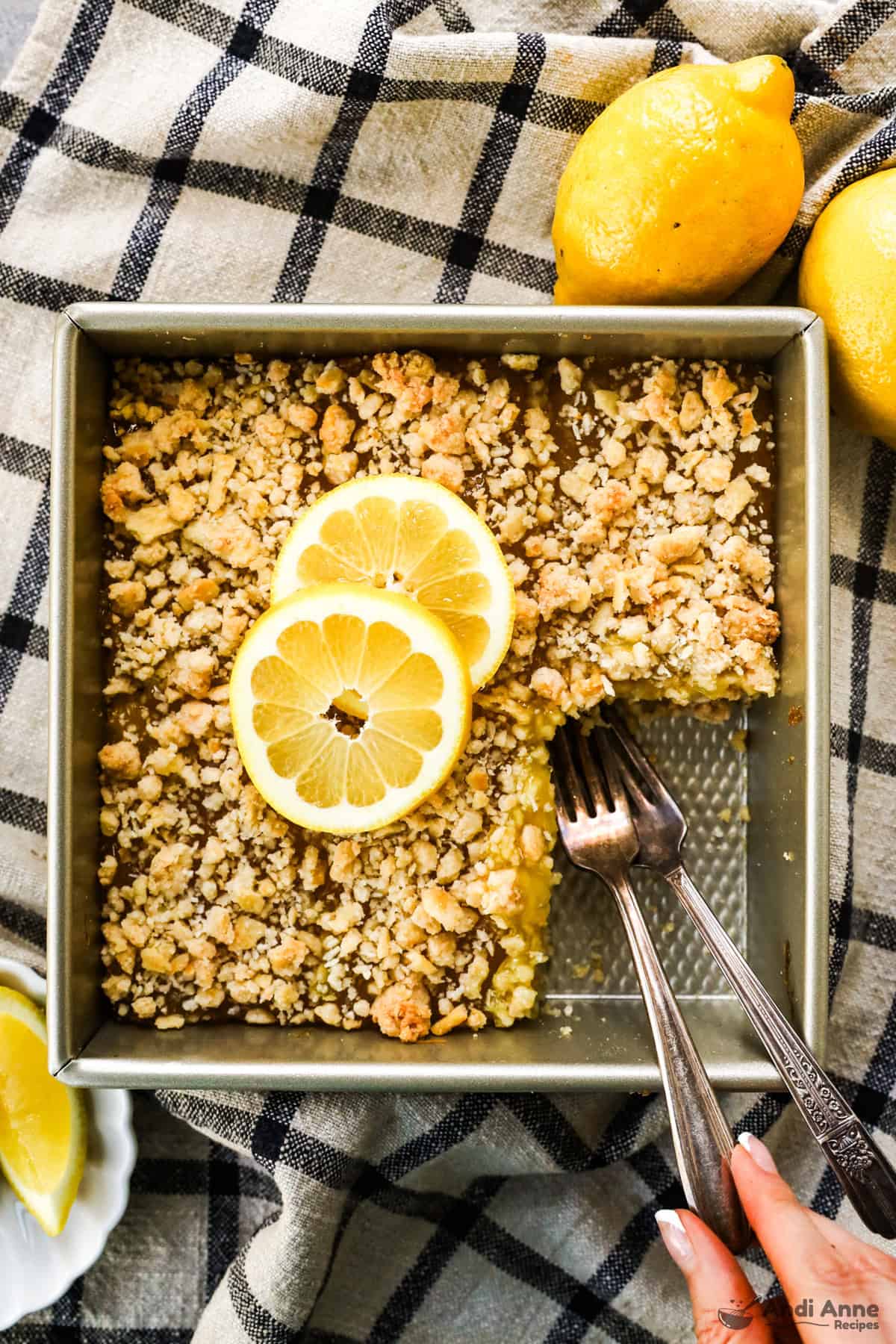 A slice is removed from the pan of lemon bars with two forks, and two lemon slices are placed on top.