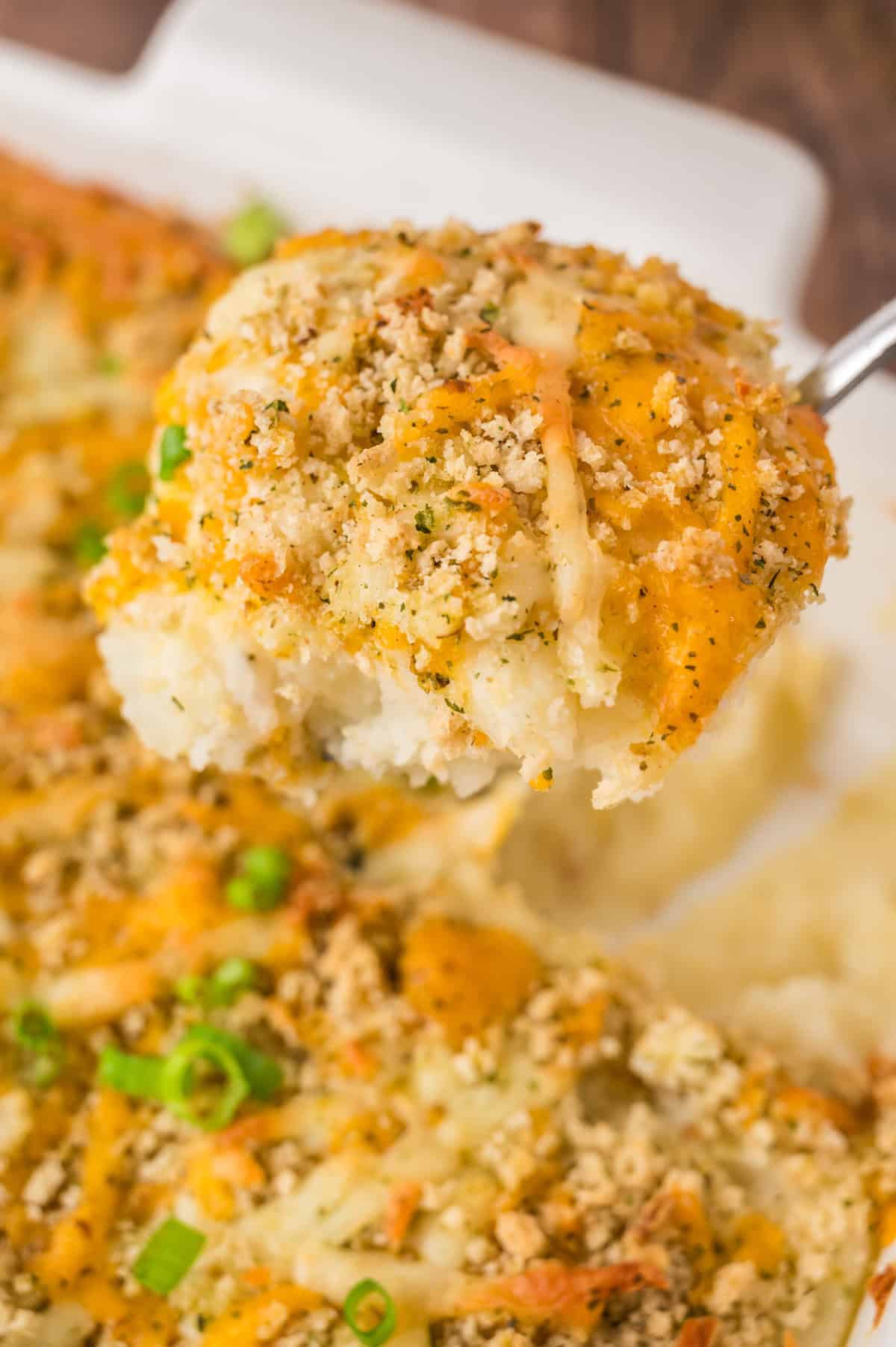 A spoon is lifting a serving of mashed potato casserole topped with bread crumbs and cheese.