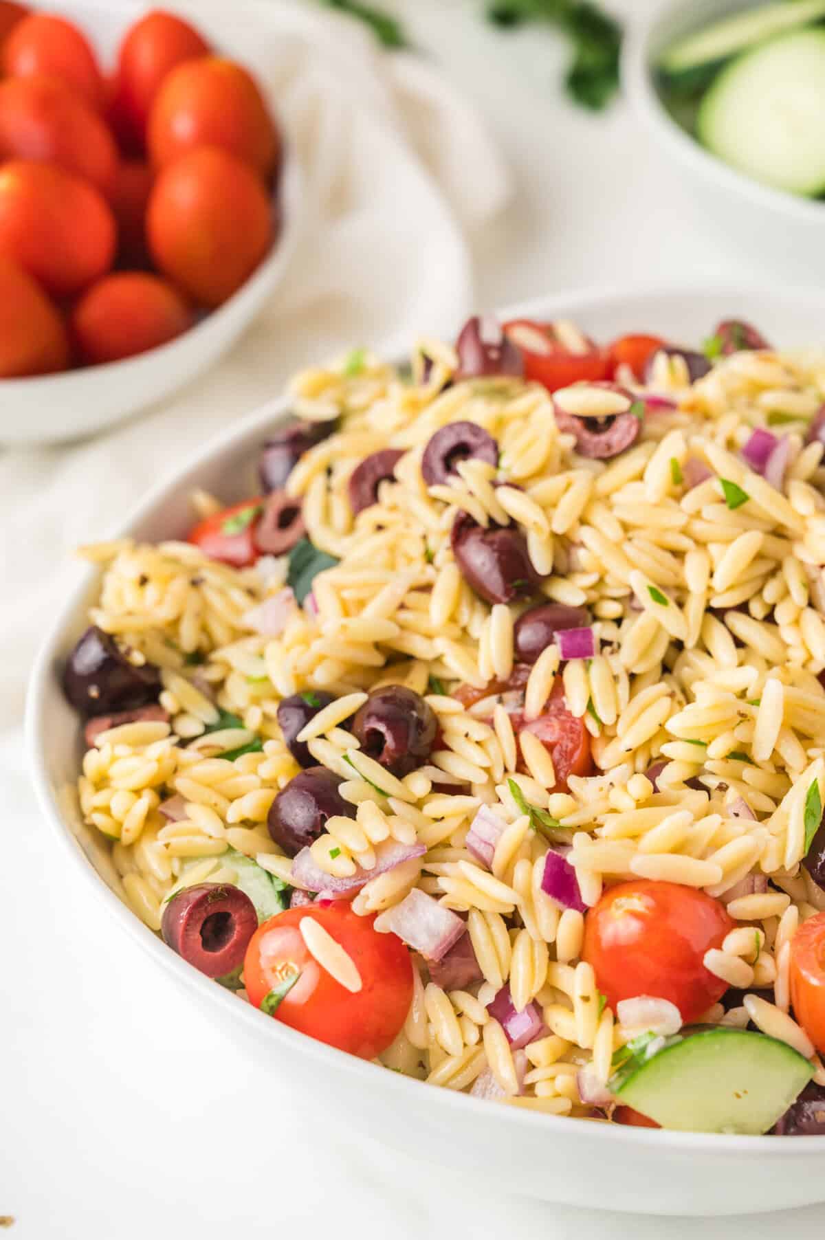 A large white bowl filled with orzo salad with tomatoes, olives, cucumber, and feta cheese.