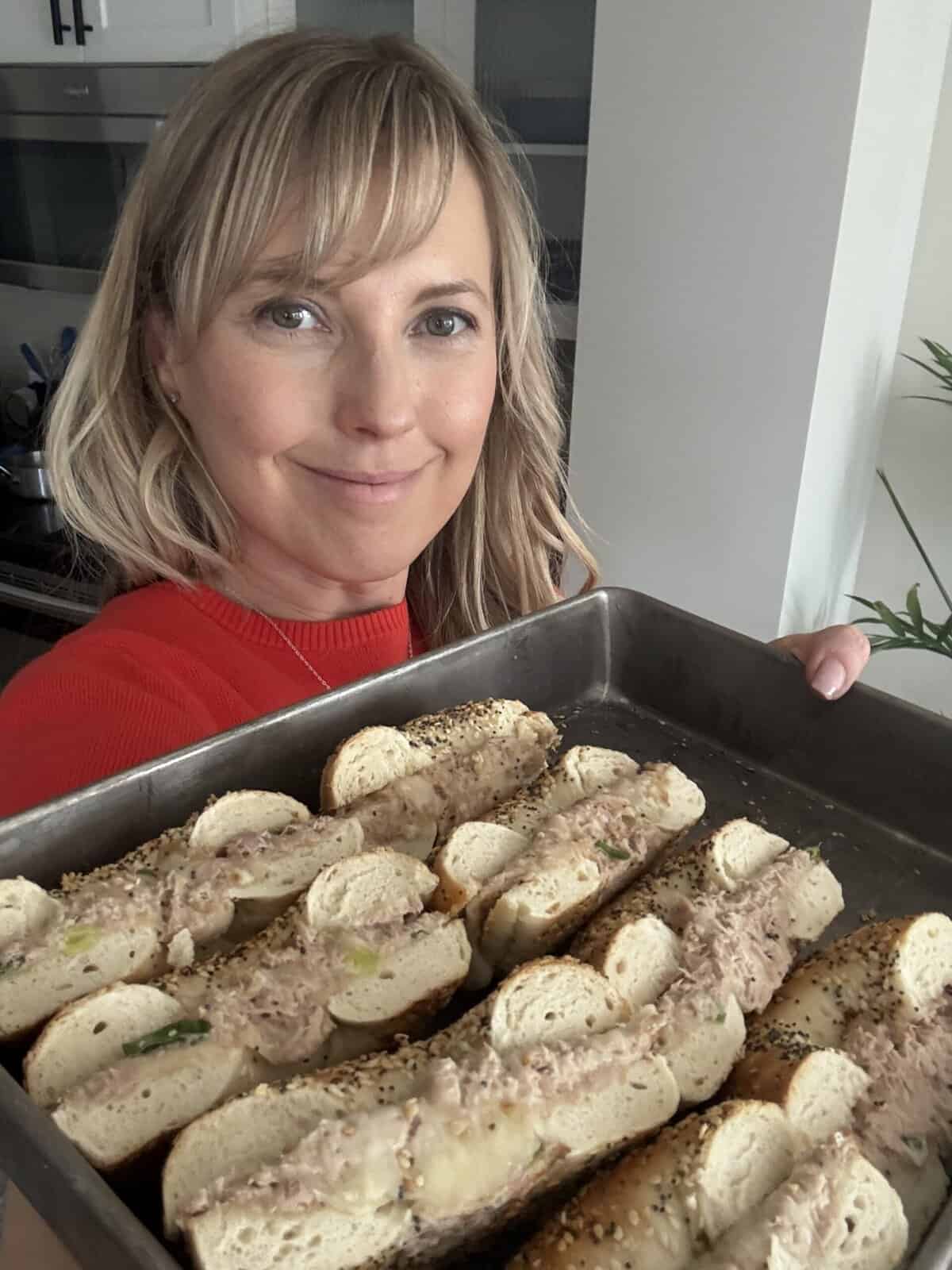 Andi is holding a sheet pan with halved bagel tuna melts.