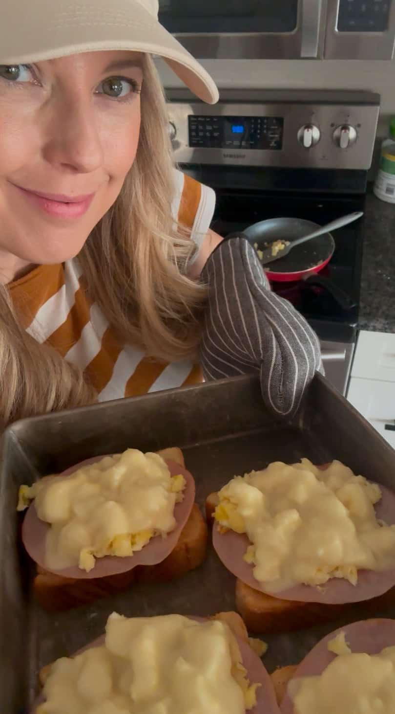 Andi is holding a baking tray with four slices of toast topped with ham, scrambled eggs, and cheese.