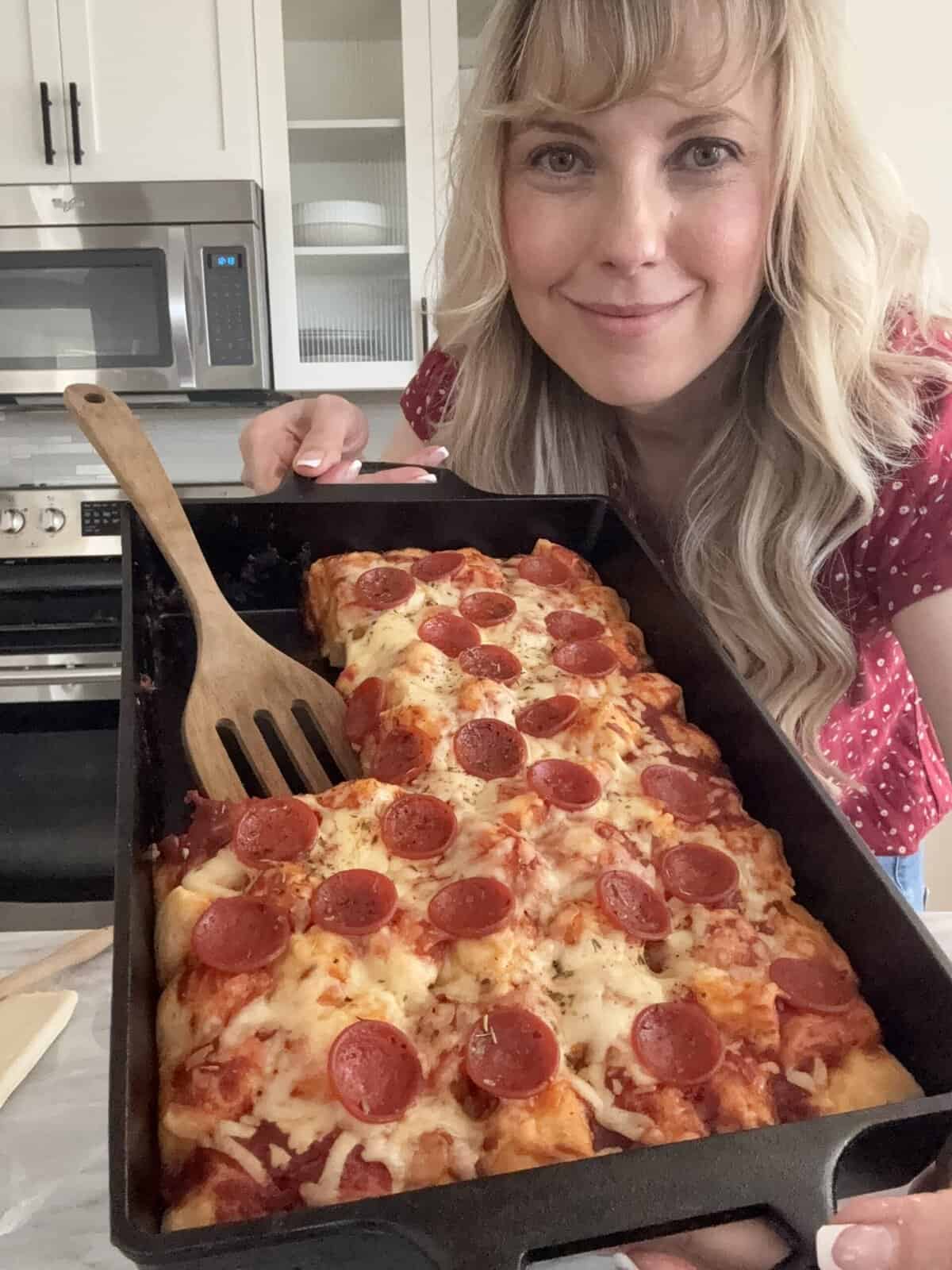 Andi Anne holding a freshly baked pizza biscuit casserole in her kitchen, showing the golden cheese and pepperoni on top.