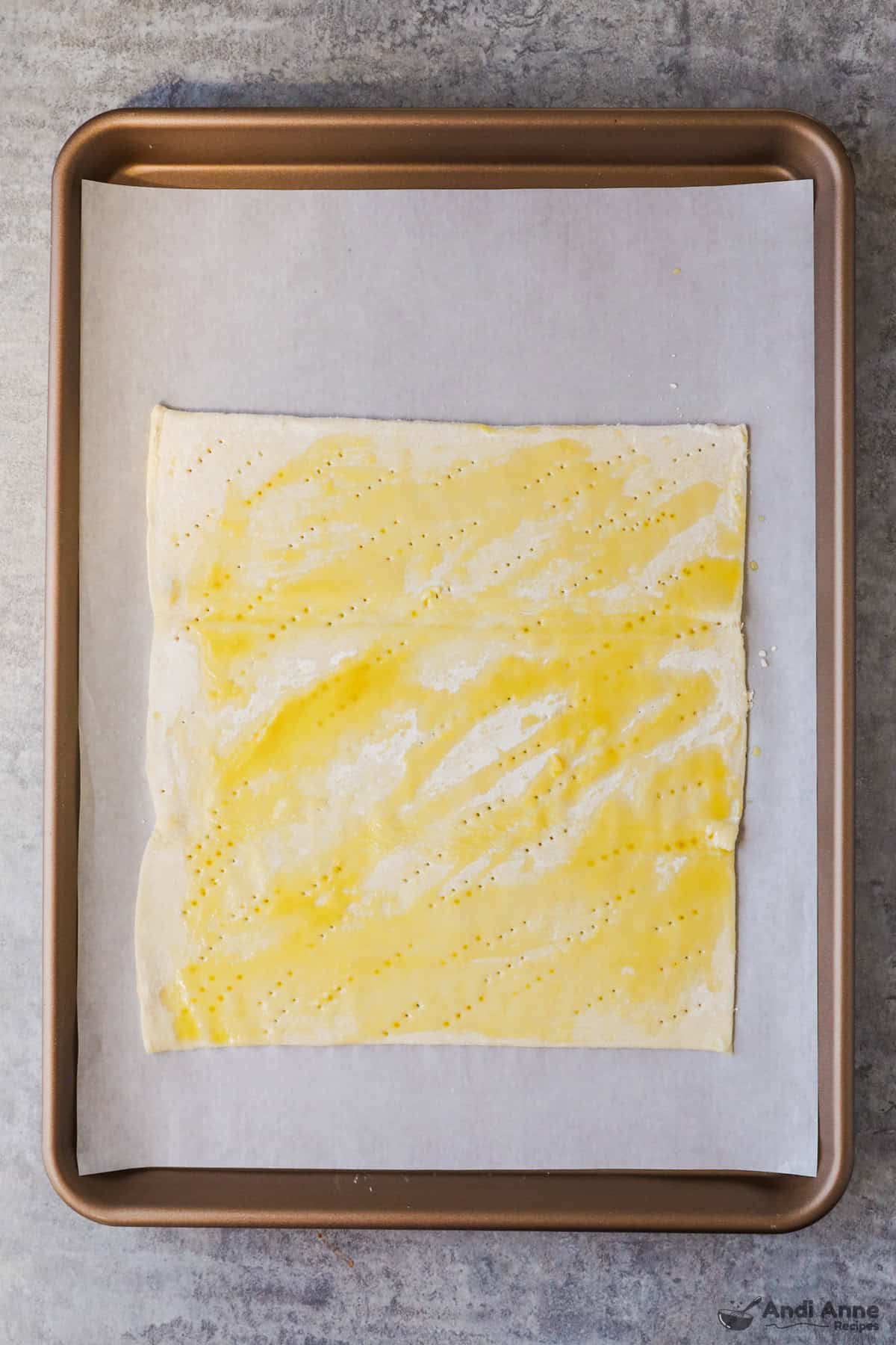 A sheet of puff pastry dough on a baking tray lined with parchment has been docked and drizzled with olive oil.