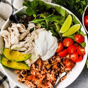 A burrito bowl with black beans, rice, tomatoes, salad greens, avocado, shredded chicken, and topped with sour cream.