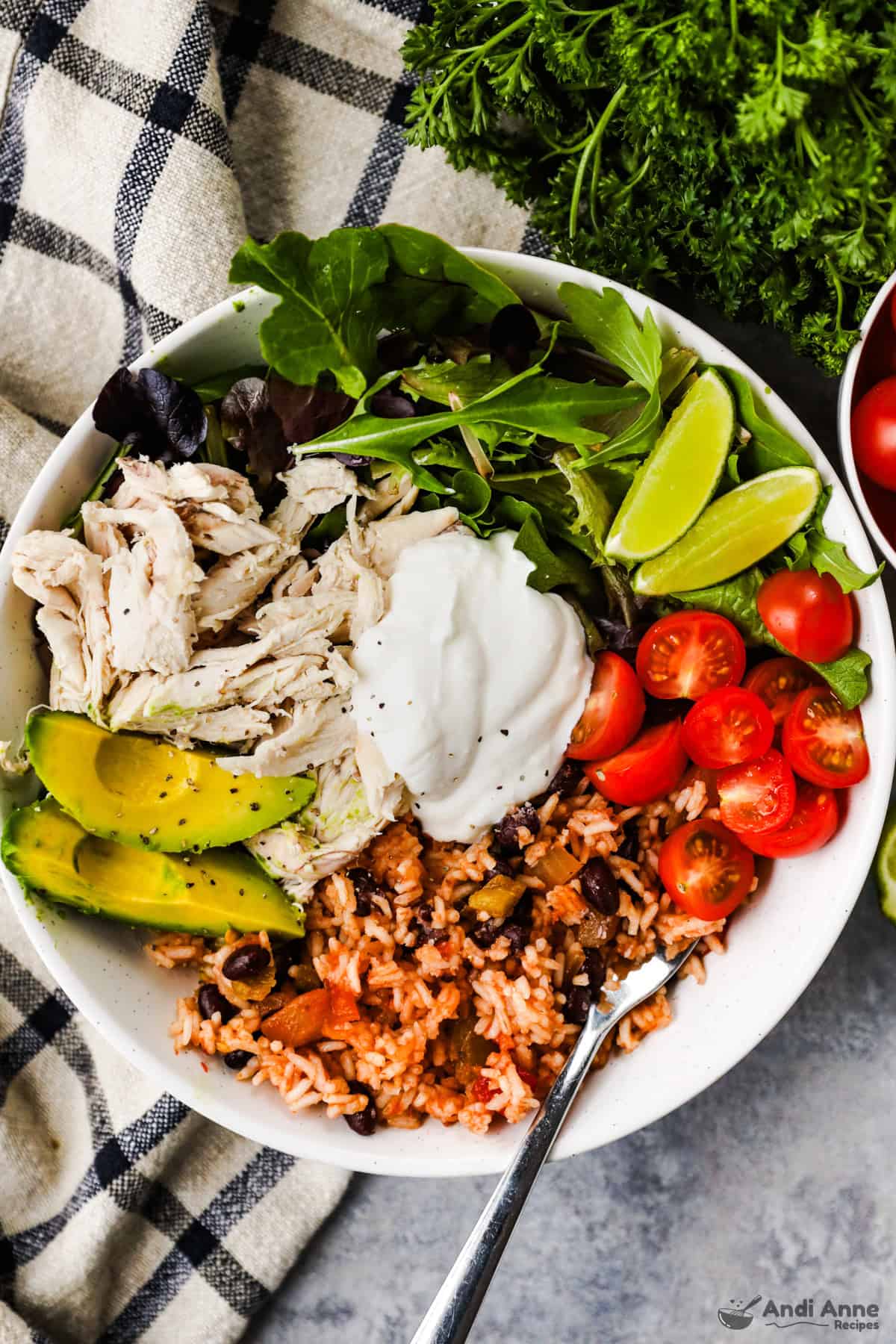 A burrito bowl with black beans, rice, tomatoes, salad greens, avocado, shredded chicken, and topped with sour cream.