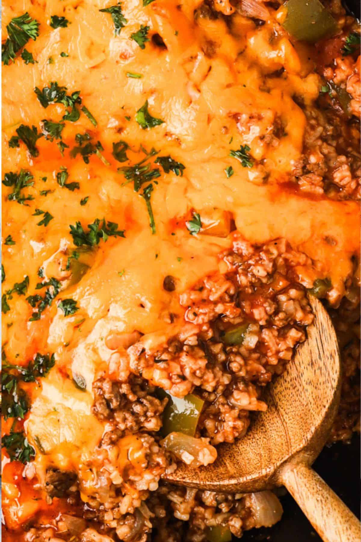 A wooden spoon lifts ground beef, rice, bell peppers, tomato sauce, and melted cheese from a skillet.
