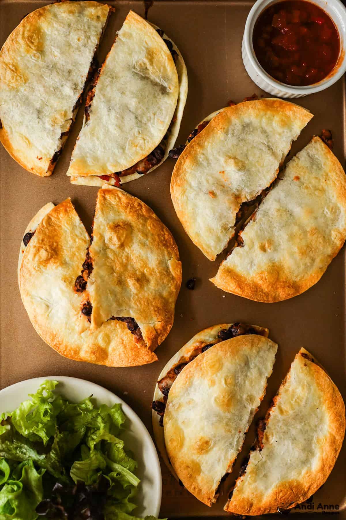 The crispy black bean quesadillas are cut in half on a baking sheet with a side dish of salsa and leafy salad.