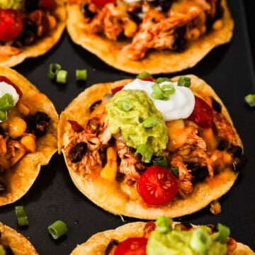 Crispy corn tostadas topped with rotisserie chicken, beans, corn, salsa, sour cream, guacamole, and green onions.