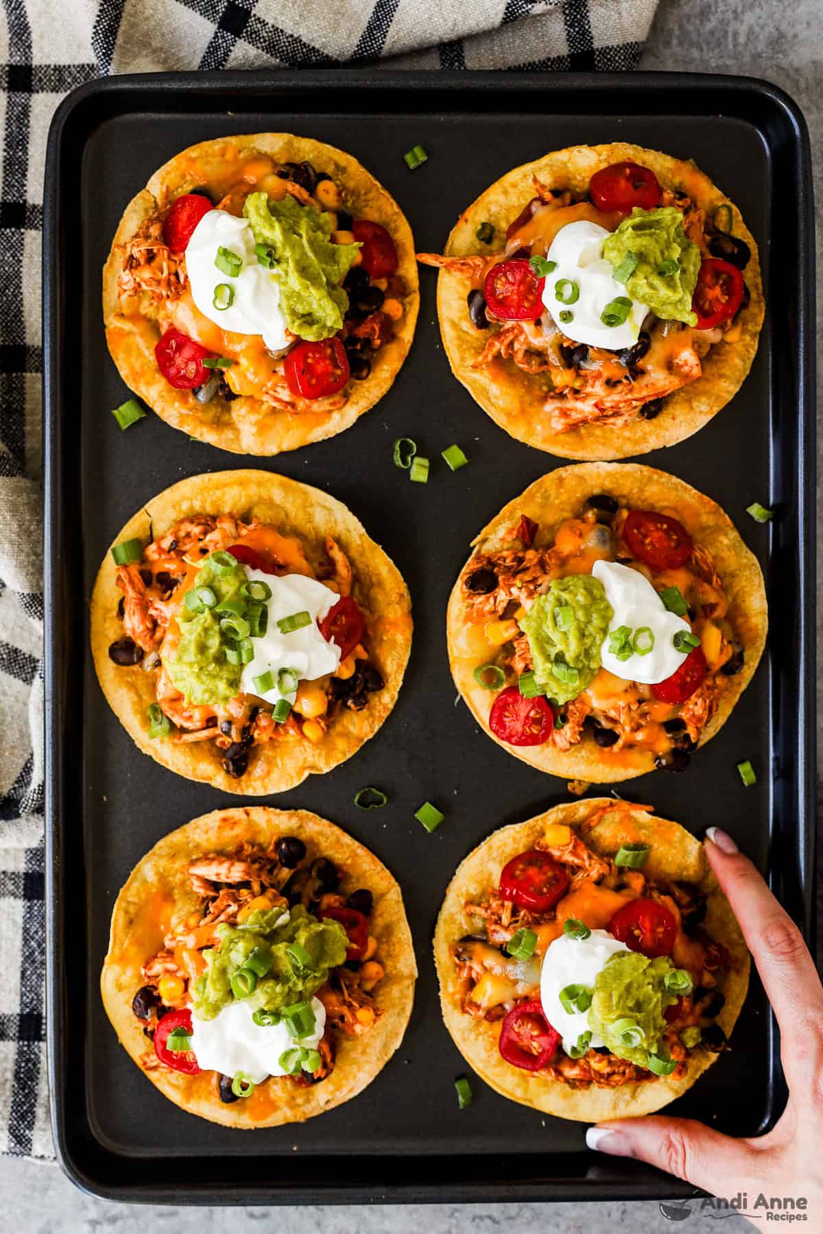 A baking tray with crispy corn tostadas topped with rotisserie chicken, beans, corn, salsa, melted cheese, sour cream, guacamole, and green onions.