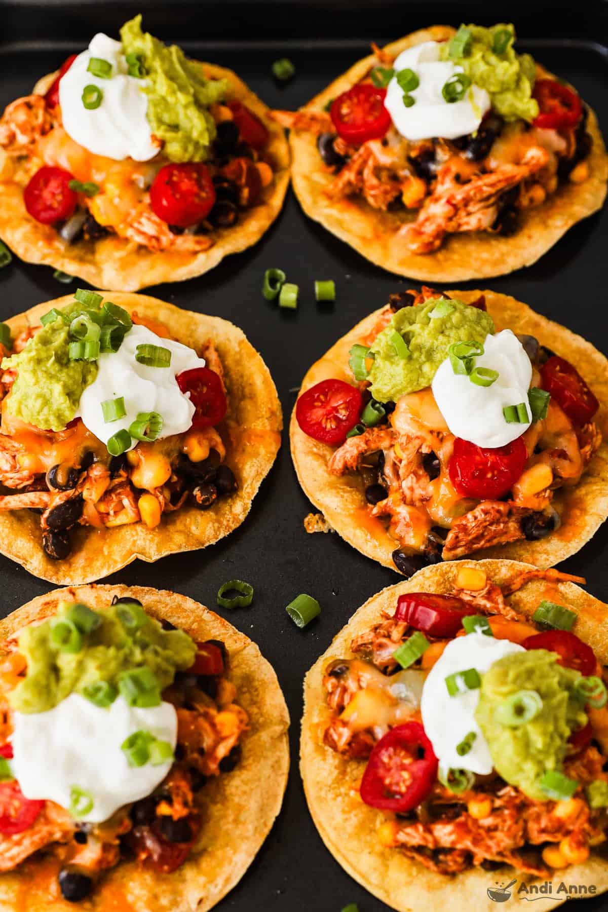A baking tray of crispy corn tostadas topped with rotisserie chicken, beans, corn, salsa, sour cream, guacamole, and green onions.