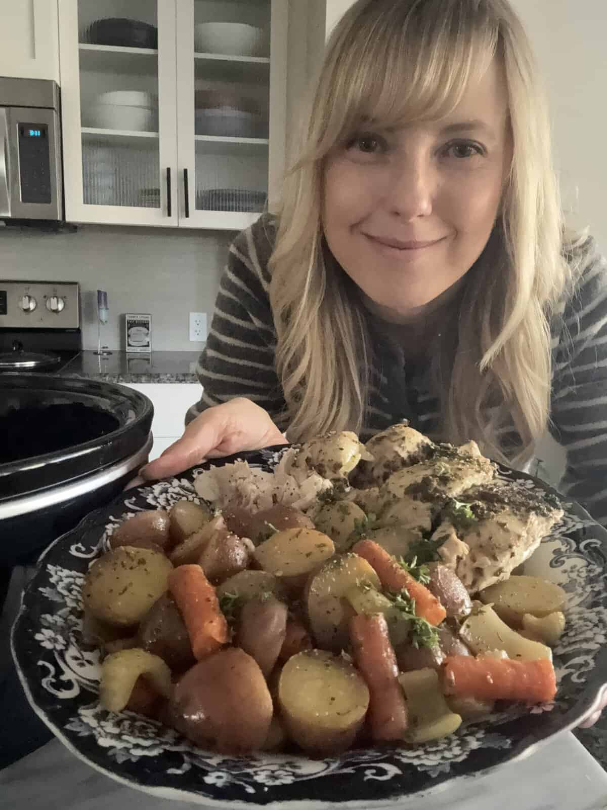 Andi is holding a serving platter with slow cooker roasted chicken and vegetables.