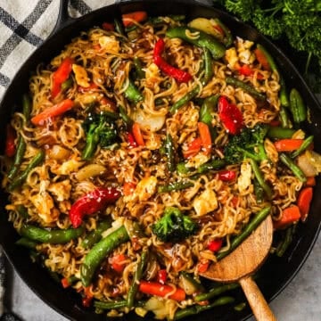 A skillet with ramen noodles, snap peas, bell peppers, carrots, and green beans in a teriyaki sauce topped with sesame seeds.