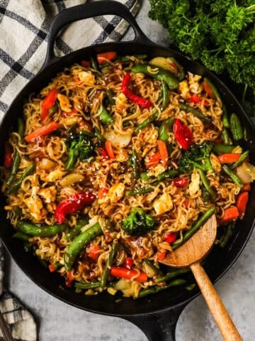 A skillet with ramen noodles, snap peas, bell peppers, carrots, and green beans in a teriyaki sauce topped with sesame seeds.