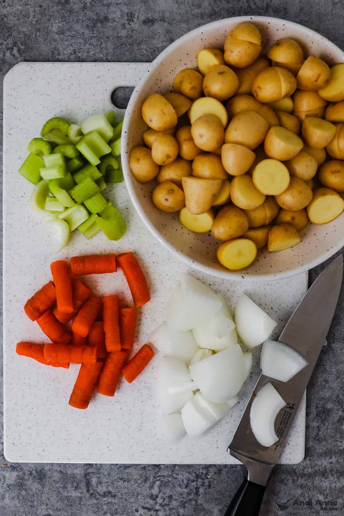 Halved baby potatoes, carrots cut into chunks, onion cut into chunks, and chopped celery on a cutting board with a knife.