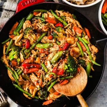 A skillet with shredded rotisserie chicken, carrots, green beans, bell peppers, and snap peas in stir fry sauce, and topped with sesame seeds.