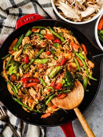 A skillet with shredded rotisserie chicken, carrots, green beans, bell peppers, and snap peas in stir fry sauce, and topped with sesame seeds.