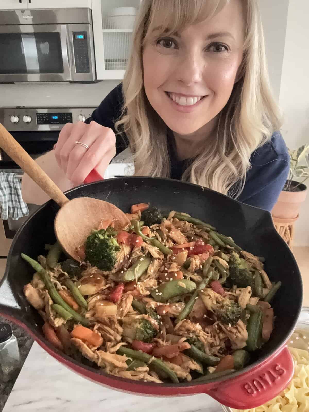Andi is holding a skillet with shredded rotisserie chicken, carrots, green beans, bell peppers, and snap peas in stir fry sauce, and topped with sesame seeds.