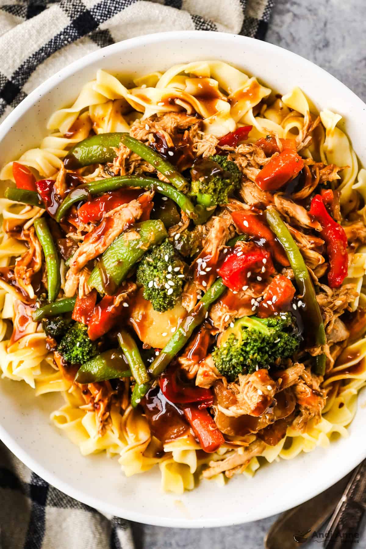 A skillet with shredded rotisserie chicken, carrots, green beans, bell peppers, and snap peas in stir fry sauce, topped with sesame seeds on a bed of egg noodles.