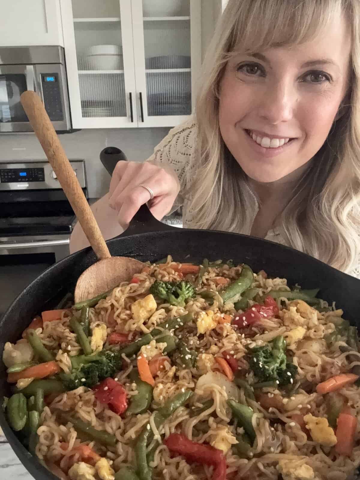 Andi is holding a skillet with ramen noodles, snap peas, bell peppers, carrots, and green beans in a teriyaki sauce topped with sesame seeds.
