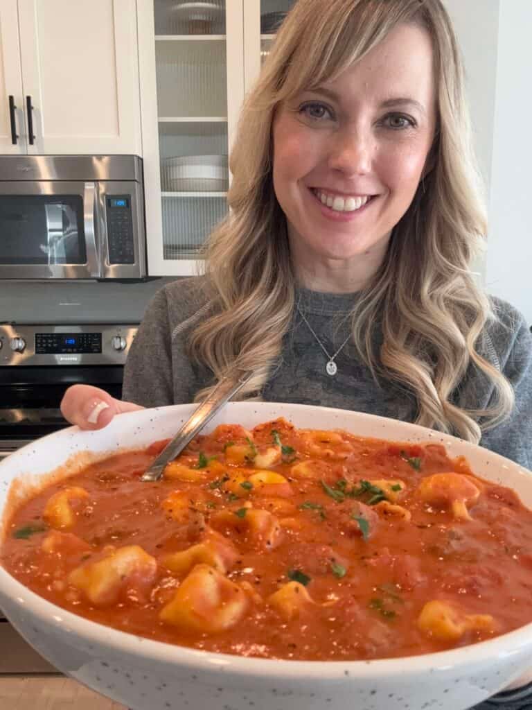 Andi is holding a large bowl of creamy tomato tortellini soup.