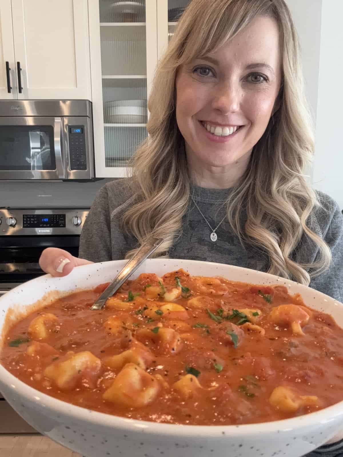 Andi is holding a large bowl of creamy tomato tortellini soup.