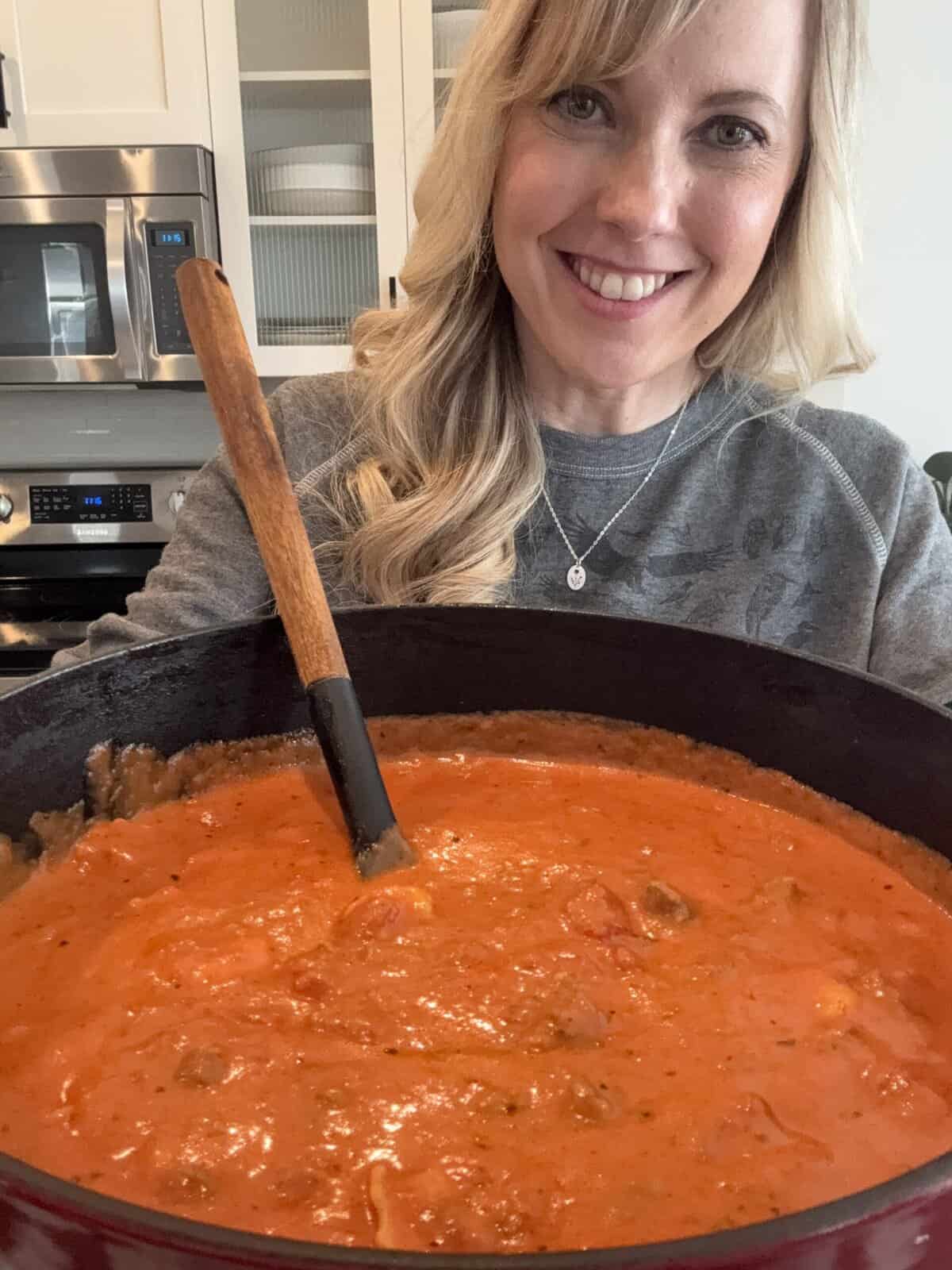 Andi is holding a large pot of creamy tomato tortellini soup with Italian sausage.