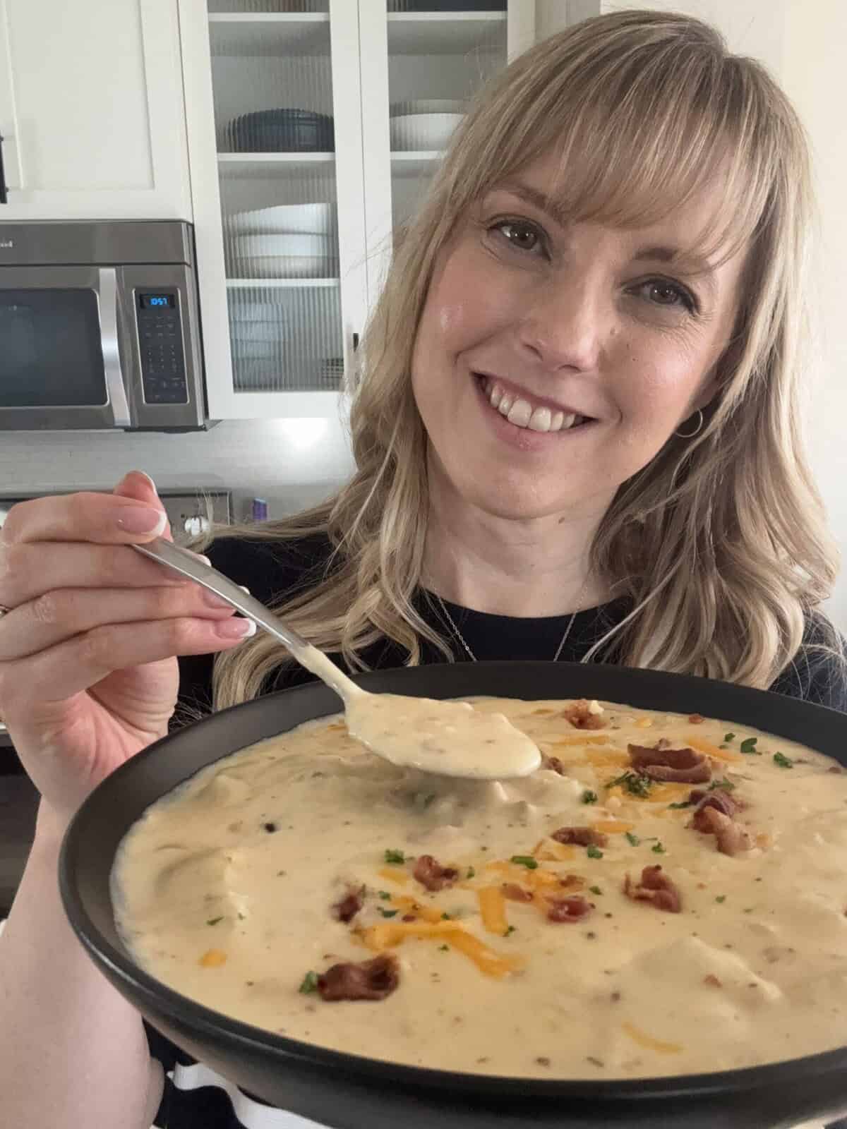 Andi is holding a bowl of loaded baked potato soup topped with cheddar cheese, bacon, and chives.