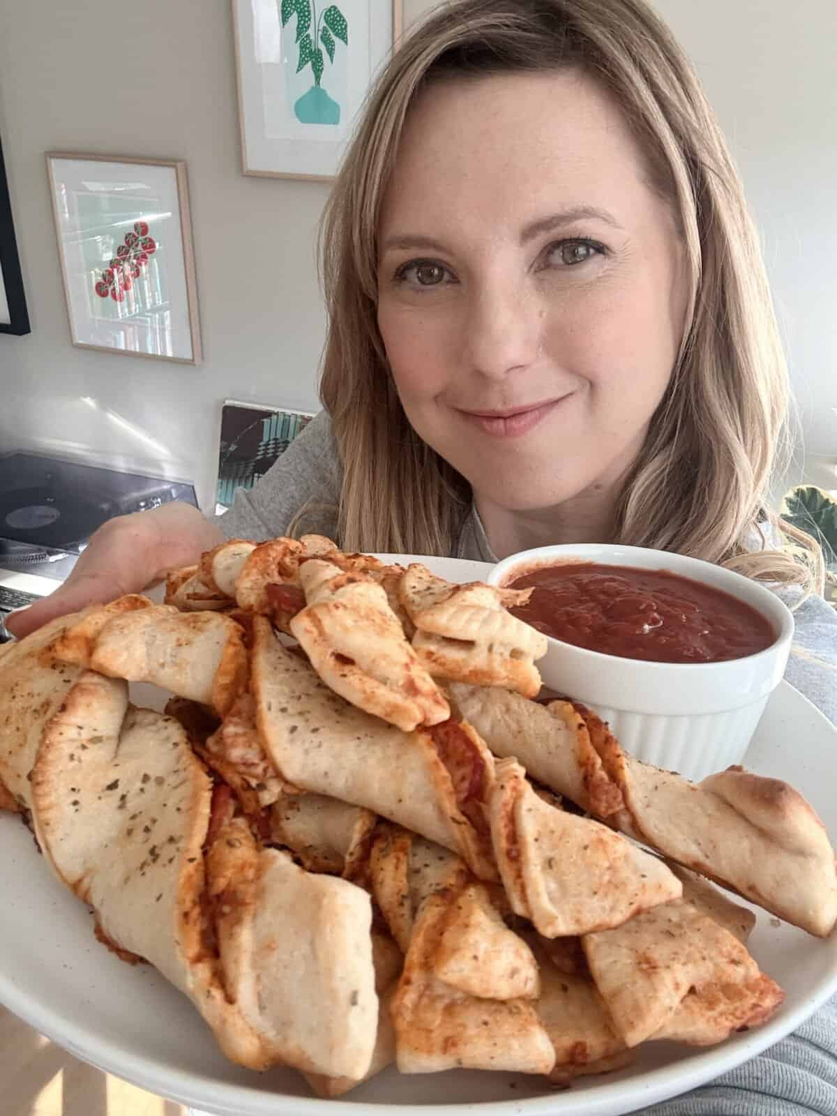 Andi is holding a plate of twisted pizza dough breadsticks with a side bowl of marinara sauce.