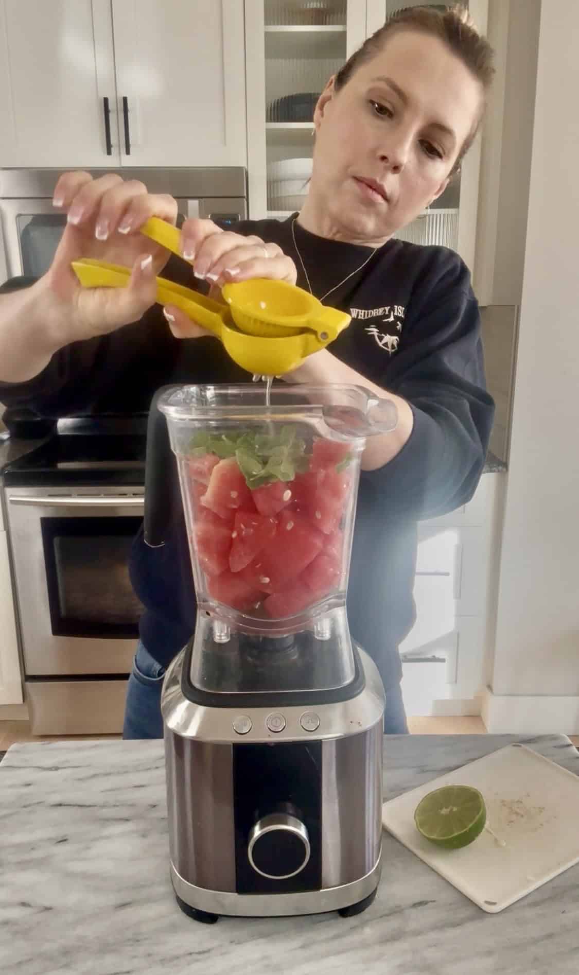 Andi is using a citrus juicer to squeeze lime juice over the cubed watermelon and mint leaves that are in the blender.