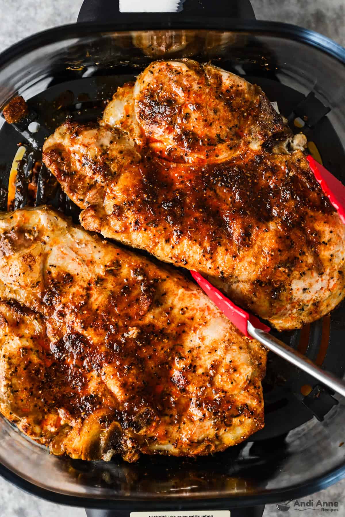 Tongs are lifting a cooked pork chop out of the air fryer.