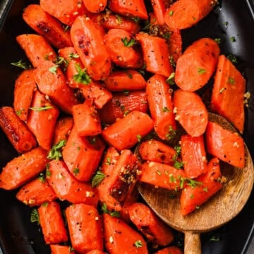 Roasted carrots with caramelized edges and garlic bits on a serving dish with a wooden spoon.