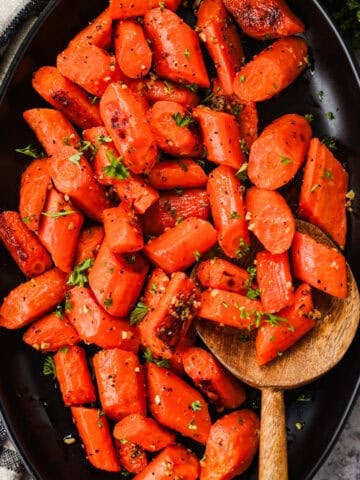 Roasted carrots with caramelized edges and garlic bits on a serving dish with a wooden spoon.