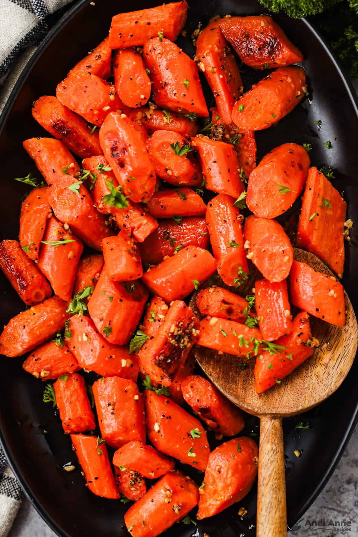 Roasted carrots with caramelized edges and garlic bits on a serving dish with a wooden spoon.