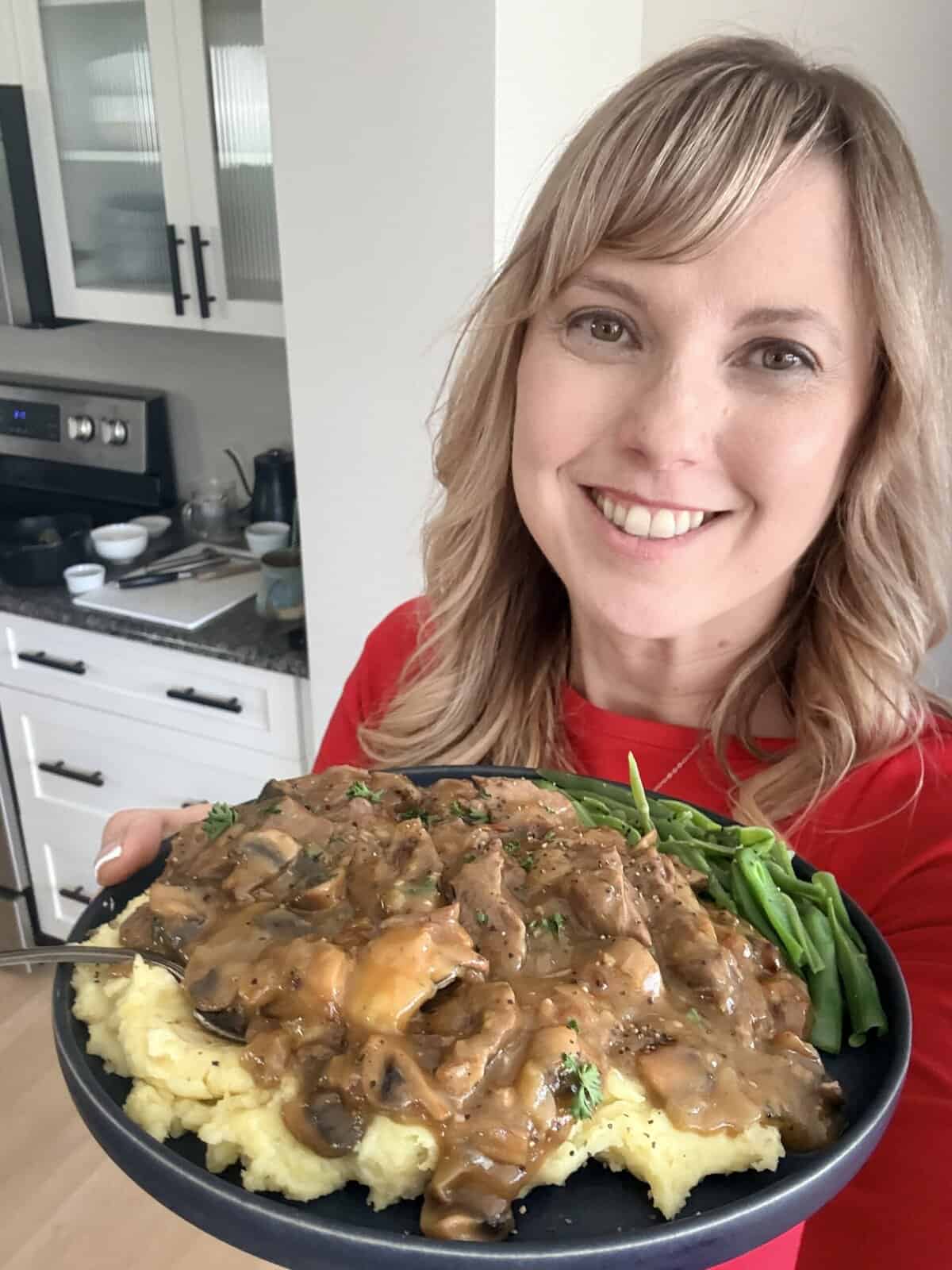 Andi is holding a plate of round steak and gravy over mashed potatoes with a side of green beans.