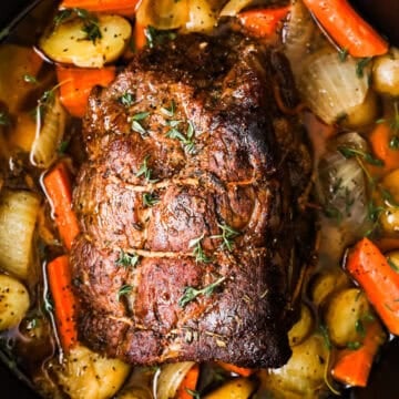 A beef roast in a Dutch oven on a bed of baby potatoes, chunks of carrot, onions, and herbs.