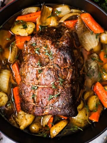 A beef roast in a Dutch oven on a bed of baby potatoes, chunks of carrot, onions, and herbs.
