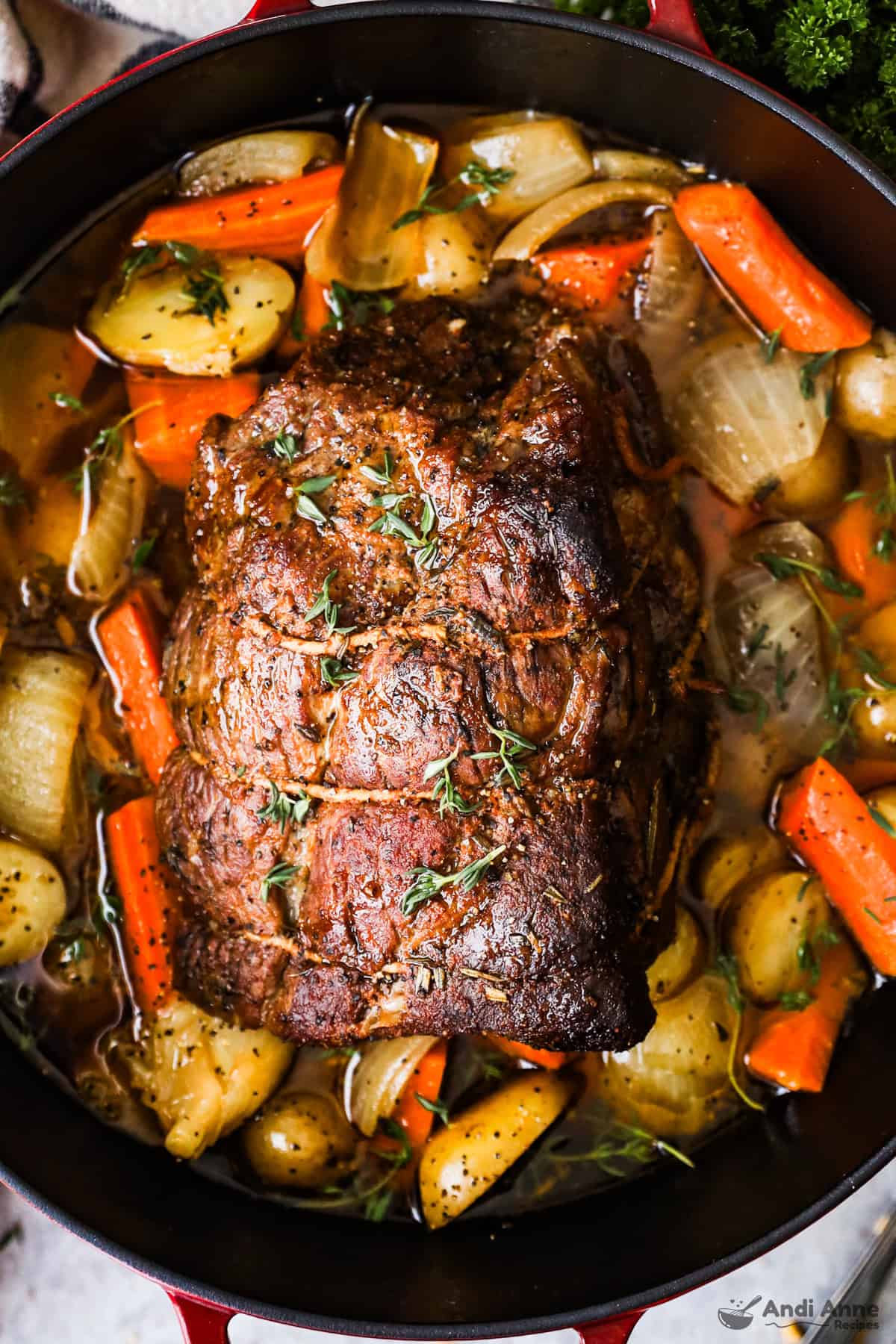 A beef roast in a Dutch oven on a bed of baby potatoes, chunks of carrot, onions, and herbs.
