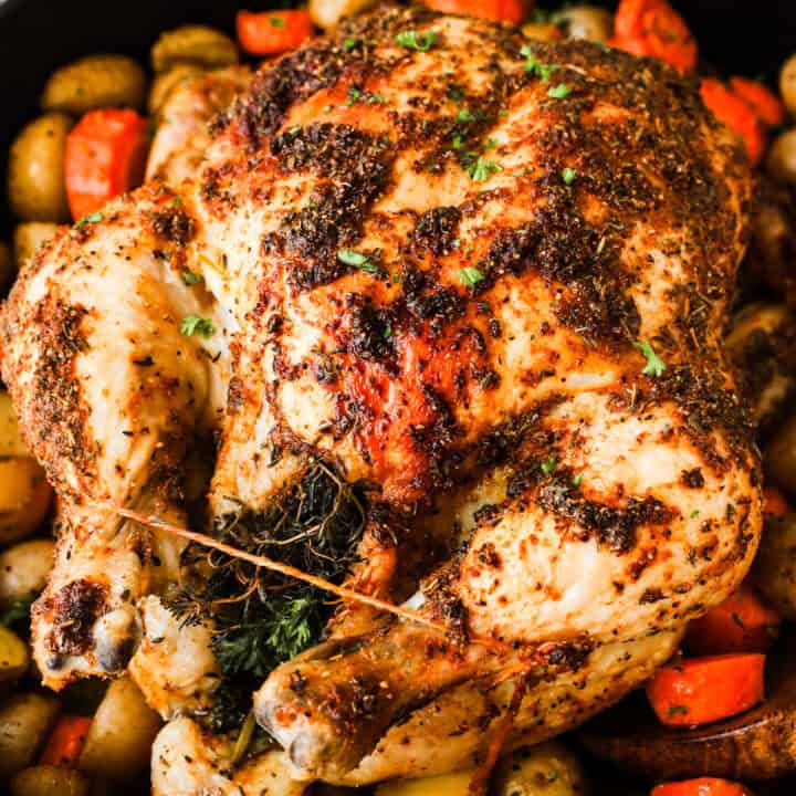 A whole roasted chicken on a bed of baby potatoes and carrot pieces in a Dutch oven.